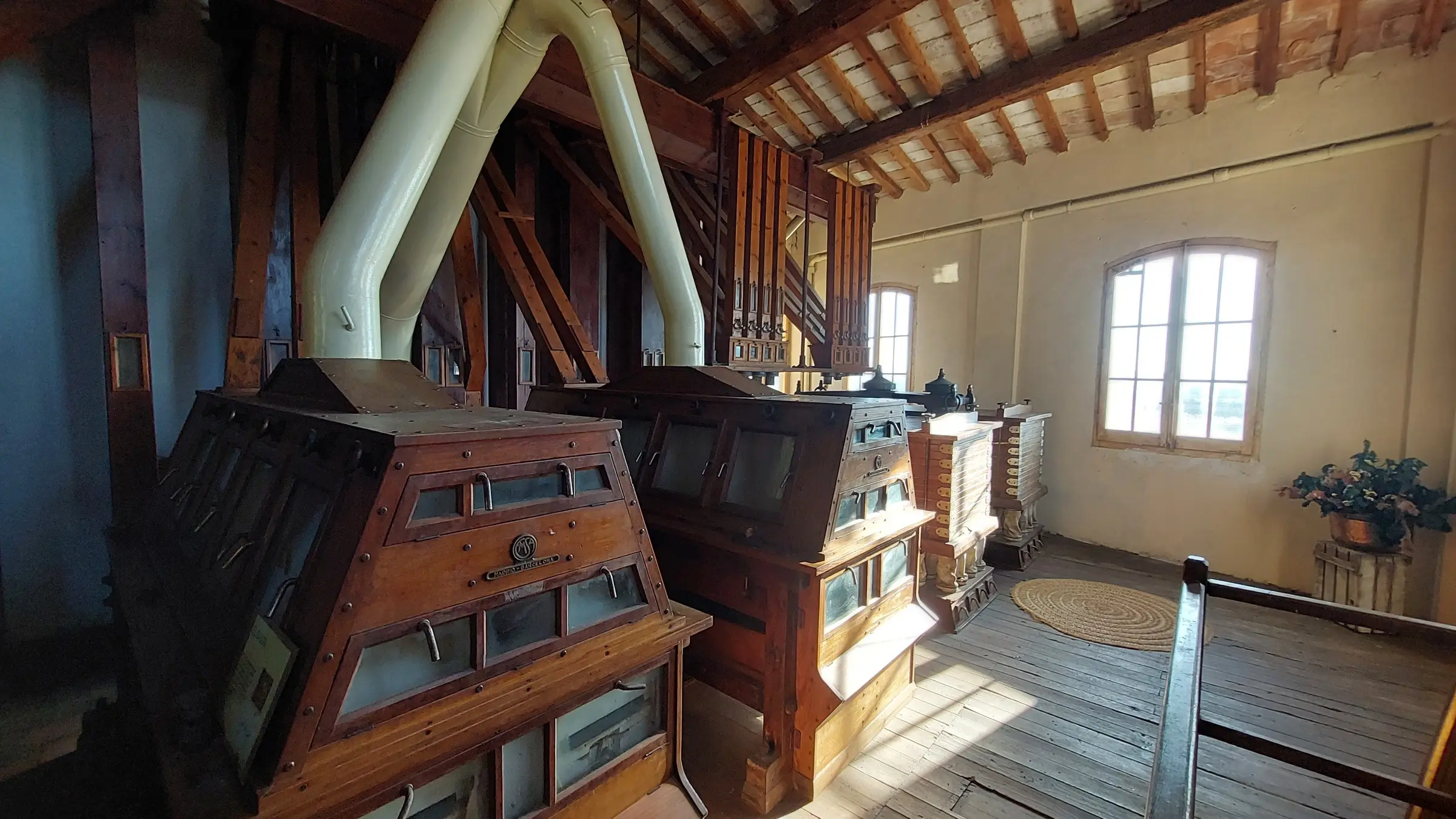 La maquinaria de madera de la antigua harinera, con sus grandes cajas y un tubo blanco, a la luz de una ventana