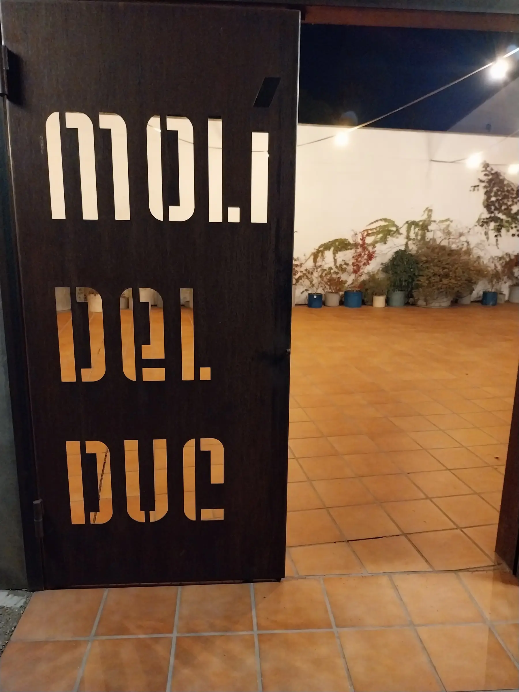 La puerta de entrada, de chapa oscura, con las letras «Molí del Duc» recortadas y la luz del interior detrás